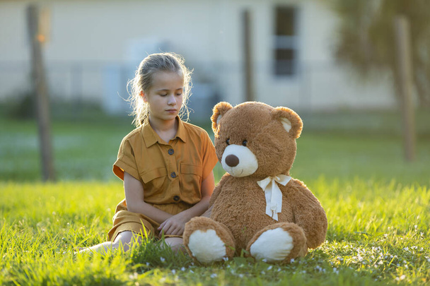 Unhappy child girl spending time with her teddy bear friend outdoors on sunny summer backyard. Concept of loneliness at preteen age and friendship problems. - Photo, Image