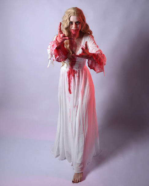  Ganztägiges Porträt einer furchterregenden Vampir-Zombie-Braut, die ein elegantes Halloween-Fantasiekostüm mit blutigem roten Farbspritzer trägt. Stehende Gehpose. Isoliert auf weißem Studiohintergrund  - Foto, Bild