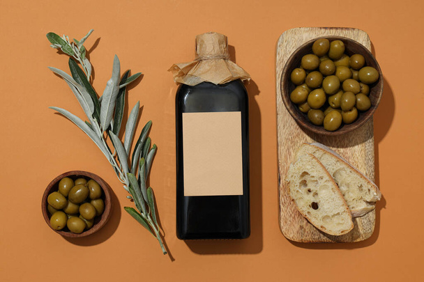 Пляшка оливкової олії, миски з оливками та скибочки хліба на помаранчевому фоні, вид зверху - Фото, зображення