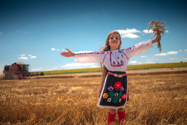 Jovem com traje tradicional de folclore búlgaro no campo de trigo agrícola durante a época de colheita com máquina de combinar industrial - Foto, Imagem