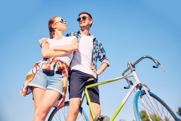 Bottom view photo of stylishly dressed couple with bicycle in summer sunlight in park. Two friends looking with love each other walking near river with cycles. Relationship leisure time concept. - Photo, Image
