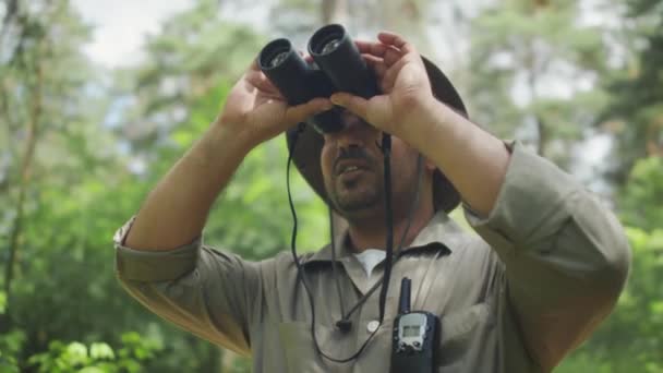 Foto de arco mediano de guardabosques macho de mediana edad con camisa caqui y sombrero de sol de pie en el bosque verde en el día de verano, mirando aves o animales a través de binoculares, y haciendo comentarios a su colega - Metraje, vídeo