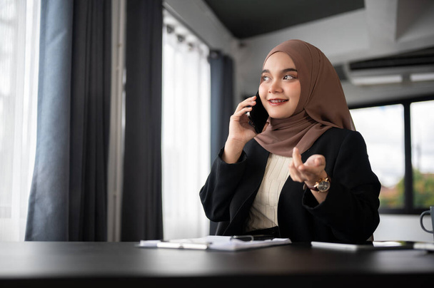 Egy magabiztos és profi felnőtt ázsiai muszlim üzletasszony vagy női főnök egy hidzsábban telefonon beszél üzleti ügyfelével az asztalánál egy modern magánirodájában.. - Fotó, kép