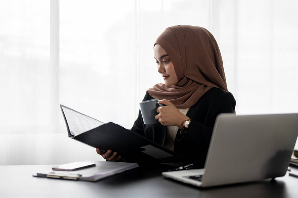Een professionele Aziatische moslim zakenvrouw of vrouwelijke manager richt zich op het onderzoeken van zakelijke rapporten in een map tijdens het drinken van haar ochtendkoffie, werken aan haar bureau in haar eigen kantoor. - Foto, afbeelding