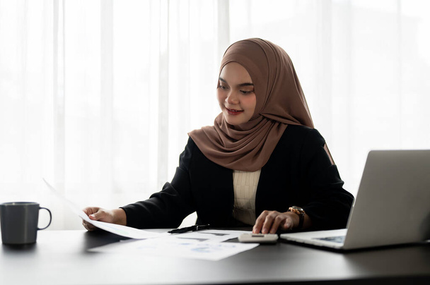 Une femme d'affaires musulmane asiatique professionnelle ou une femme gestionnaire dans un hijab travaille sur des rapports financiers, examinant les données sur les rapports à son bureau dans un bureau moderne. - Photo, image