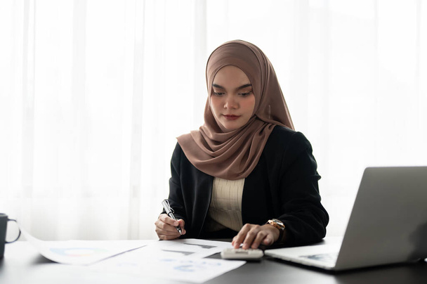 Μια επαγγελματίας και στοχαστική Ασιάτισσα μουσουλμάνα επιχειρηματίας ή γυναίκα διευθύντρια με μαντίλα επικεντρώνεται στην εργασία σε οικονομικές εκθέσεις, την εξέταση δεδομένων σχετικά με τις εκθέσεις στο γραφείο της σε ένα σύγχρονο γραφείο. - Φωτογραφία, εικόνα
