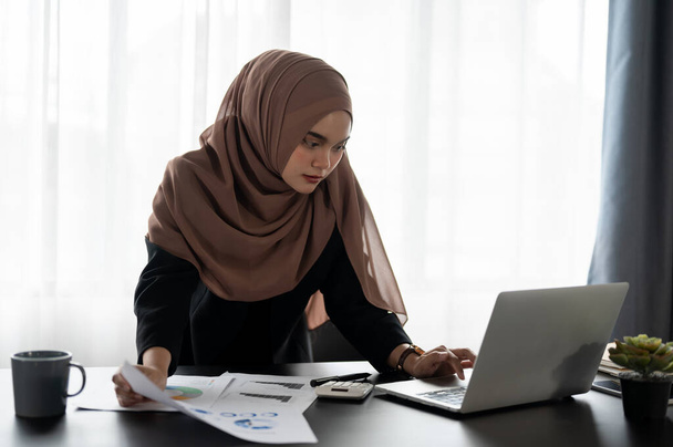 Una donna d'affari musulmana asiatica professionale e concentrata con indosso un hijab sta lavorando al suo progetto finanziario, analizzando i dati finanziari aziendali e pianificando un progetto di investimento alla sua scrivania.. - Foto, immagini