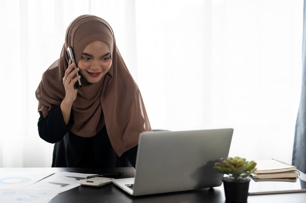 Ammattimainen ja luottavainen aasialainen muslimi liikenainen tai naispuolinen taloudellinen avustaja puhuu puhelimessa asiakkaansa kanssa, kun tarkastellaan hänen kannettavan tietokoneensa yksityiskohtia toimistossaan.. - Valokuva, kuva