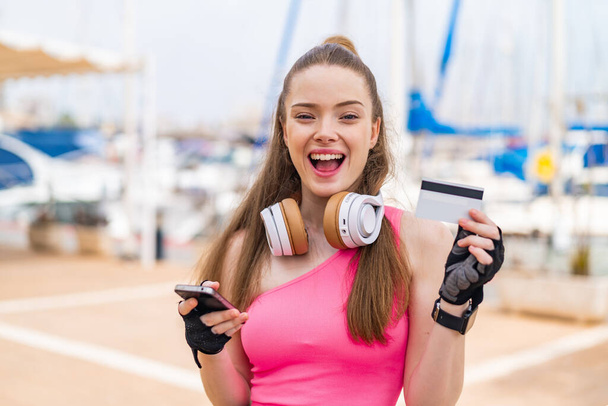 Молода красива спортивна дівчина на відкритому повітрі купує з мобільним телефоном і тримає кредитну картку з здивованим виразом - Фото, зображення