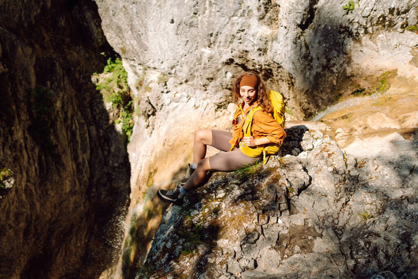 Smiling female traveler with a yellow backpack, dressed in hiking clothes, enjoys a waterfall near a mountain river. Travel, trekking. Nature concept. - Zdjęcie, obraz