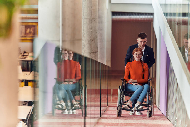 Ein Firmendirektor unterstützt seine Geschäftspartnerin im Rollstuhl und hilft ihr, zu ihrem Startup-Büro zu gelangen, wo sie mit ihrem vielfältigen Team von Kollegen zusammenarbeiten und dabei Inklusivität und Integration betonen. - Foto, Bild