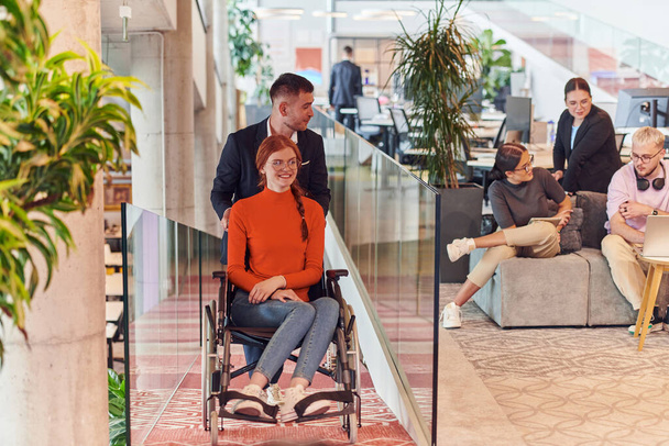 A company director assists his business colleague in a wheelchair, helping her navigate to their startup office, where they work alongside their diverse team of colleagues, emphasizing inclusivity and - 写真・画像