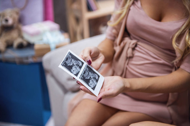 Περικομμένη εικόνα όμορφη έγκυος γυναίκα κρατώντας ένα υπερηχογράφημα ενώ κάθεται στον καναπέ στο σπίτι - Φωτογραφία, εικόνα