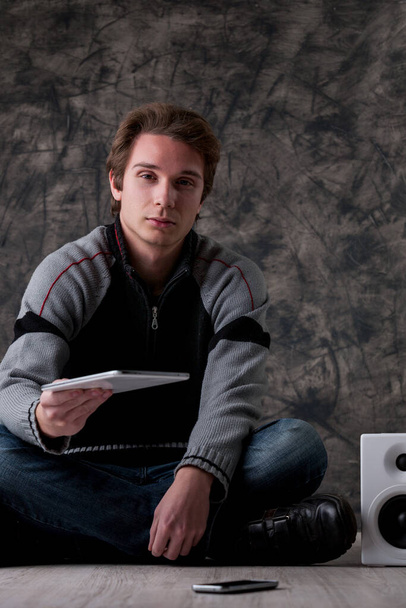 Νεαρός άνδρας με γκρι και μαύρο πουλόβερ στέκεται σε εσωτερικούς χώρους, κοντά σε λευκά ηχεία hi-fi και ένα smartphone. Επεκτείνει ένα ψηφιακό tablet, προωθώντας mobile tech, εφαρμογές, και online εργαλεία για την εργασία και την ψυχαγωγία - Φωτογραφία, εικόνα