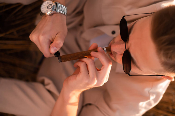 Жорстокий молодий чоловік в сонцезахисних окулярах, в бежевому костюмі на полі і курильних сигаретах - Фото, зображення