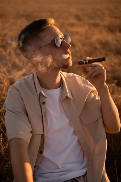 Brutální mladý muž ve slunečních brýlích, v béžovém obleku na hřišti a kouřící cigarety - Fotografie, Obrázek