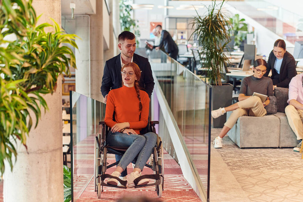 Een directeur van een bedrijf helpt zijn collega in een rolstoel, helpt haar navigeren naar hun startup office, waar ze werken samen met hun diverse team van collega 's, met de nadruk op inclusiviteit en - Foto, afbeelding