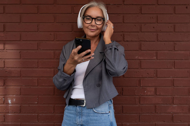 encantadora mulher madura esbelta com óculos vestidos com uma jaqueta cinza e jeans escuta música em fones de ouvido. - Foto, Imagem