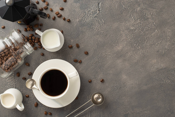 Sueño del amante del café: Vista superior de granos de café dispersos, taza de café expreso, crema y tarro de leche, cuchara de barista y hervidor de agua en una superficie gris texturizada, con espacio para su texto - Foto, Imagen