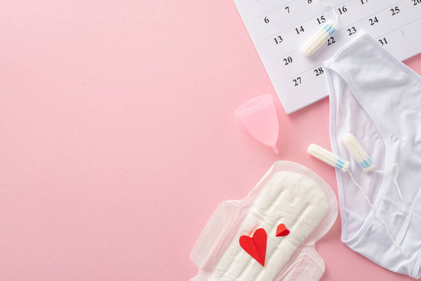 Feminine hygiene supplies like pad with red hearts, symbolizing blood, tampons, menstrual cup, underpants, calendar marking the cycle start, on a pastel pink background with room for text or branding - Photo, Image
