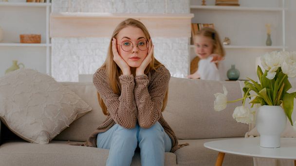 Tired upset Caucasian mother sitting on sofa at home sad frustrated stressed exhausted fatigued mom suffer headache with active noise difficult kid child baby girl daughter running around playing - Foto, imagen