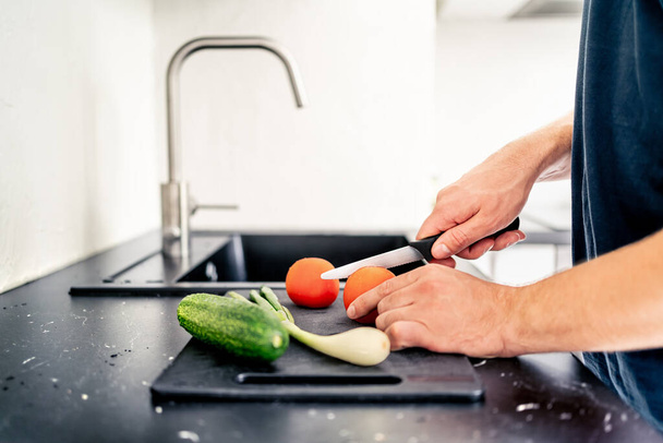 鋭いナイフでサラダを切る。 料理する男. トマトを切って野菜をスライスする. 家庭のキッチンで食べ物を準備する. 健康的なベジタリアン料理。 オーガニックビーガン成分。 ランチやディナーを作る. - 写真・画像