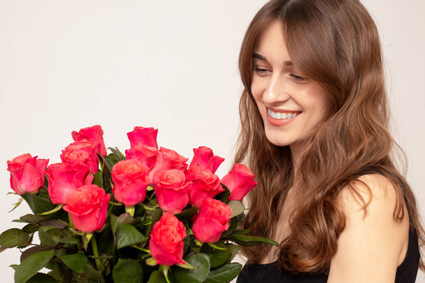 Young attractive girl with a bouquet of red roses on a white background. A happy girl with a bouquet of roses is smiling. The concept of happiness, joy and celebration - Photo, Image