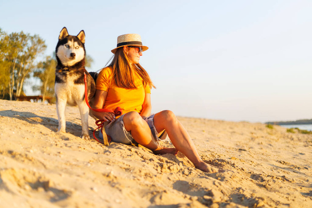 Fröhliche hübsche junge Frau mit Hut und Sonnenbrille sitzt mit ihrem Hund am Strand und genießt den Sonnenuntergang - Foto, Bild