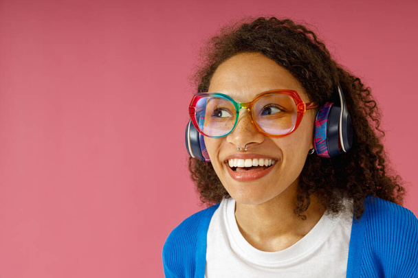 Joyful woman in headphones and glasses looking at side standing on pink studio background - Photo, Image