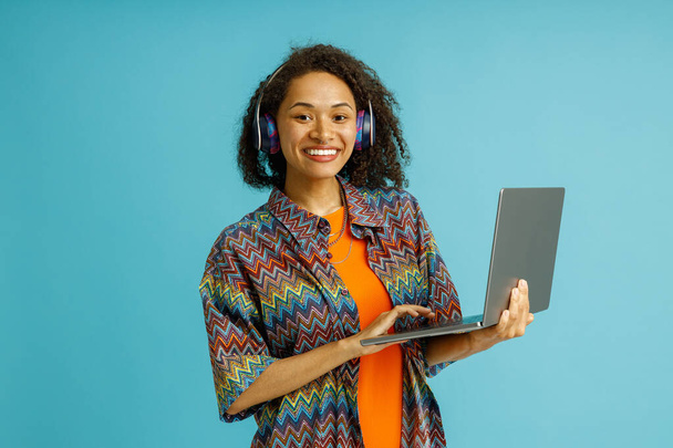 Радостная студентка в наушниках учится на ноутбуке, стоя на синем фоне студии - Фото, изображение