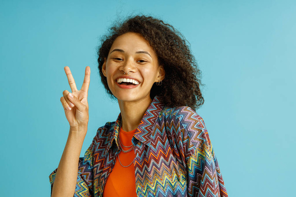Счастливая стильная женщина показывает знак мира и смотрит в камеру на голубом студийном фоне  - Фото, изображение