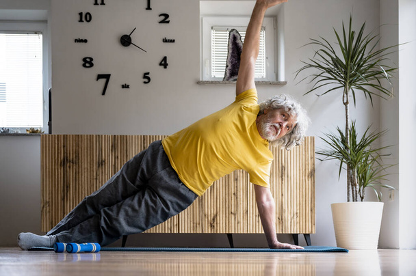 Активный старший мужчина в желтой футболке заботится о своем здоровье, тренируясь и тренируясь. Делать боковую доску положение для сильного ядра на коврике в домашней гостиной. - Фото, изображение