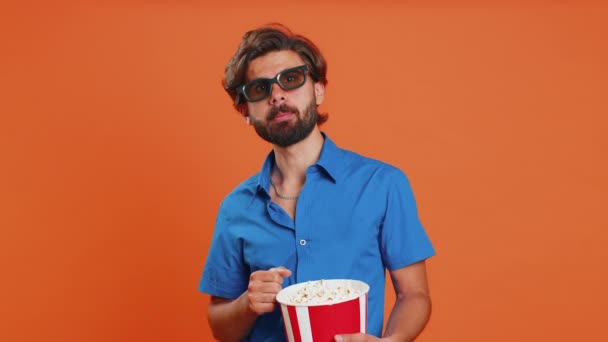 Ενθουσιασμένος νεαρός άνδρας σε 3D γυαλιά τρώει ποπ κορν και βλέποντας ενδιαφέρουσα τηλεοπτική σειρά, σπορ παιχνίδι, ταινία, online περιεχόμενο ταινία social media. Μέση Ανατολή τύπος σε πουκάμισο απομονώνονται σε πορτοκαλί στούντιο φόντο - Πλάνα, βίντεο