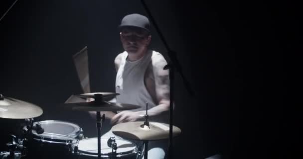 Cool joven baterista masculino con tatuajes en el cuerpo en la tapa usando palos mientras toca la batería con los ojos cerrados durante el ensayo en el estudio oscuro - Imágenes, Vídeo
