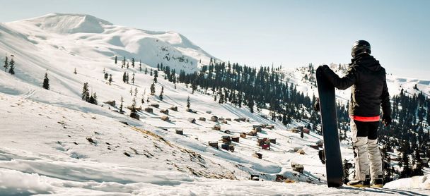 ダークブラックスポーツウェアを着た男性スノーボーダースタンドは,ジョージアのスキーホリデーの目的地ゴダルジ,コーカサス山脈の山のスキー場の風景のパノラマを楽しんでいます - 写真・画像