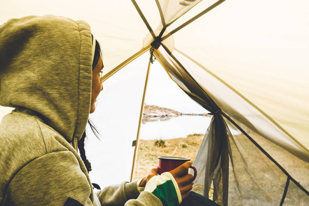 Smiley young caucasian woman hold camping mug and drink in green tent outdoors in nature with viewpoint panorama of alpine lake in autumn - Фото, зображення