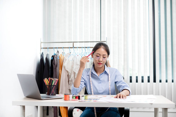 Belle jeune tailleur de mode asiatique avec des accessoires de couture textile et des croquis de designer entrepreneur sont pleins de couleurs vives sur le bureau avec un ordinateur portable pour des idées créatives en studio. - Photo, image