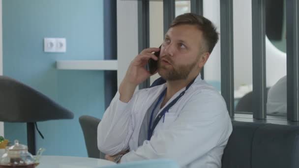 Mężczyzna-lekarz siedzi w kawiarni klinicznej, odpoczywa na przerwie. Profesjonalny lekarz rozmawia z pacjentem przez telefon, omawia wyniki badań medycznych lub leczenia. Personel medyczny w stołówce szpitalnej lub centrum medycznym. - Materiał filmowy, wideo