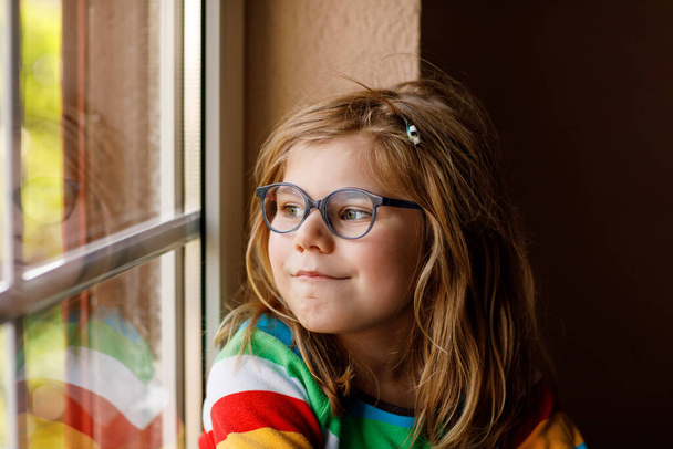 Adorable petite fille d'âge préscolaire avec des lunettes assis près de la fenêtre. Enfant réfléchi regardant dehors. Enfant solitaire - Photo, image