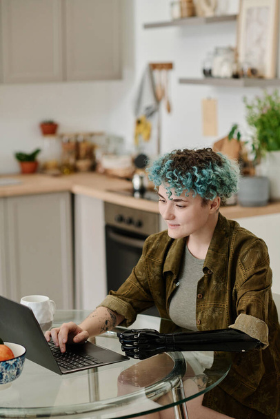 Vertical image of woman with prosthetic arm paying for her purchase online using laptop while sitting in the kitchen - Photo, Image