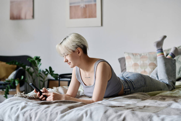 Девушка лежит на кровати в спальне и печатает сообщение на смартфоне - Фото, изображение