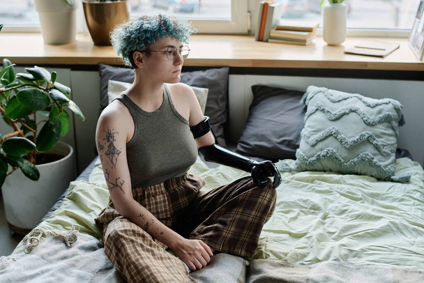 Chica con brazo protésico sentada en la cama con expresión triste y pensando en algo - Foto, Imagen
