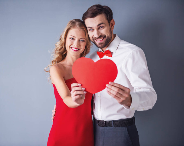 Mooie elegante meisje in de rode jurk en man in klassieke shirt en rode vlinderdas houden een papier-hart, camera kijken en glimlachend, op grijze achtergrond - Foto, afbeelding