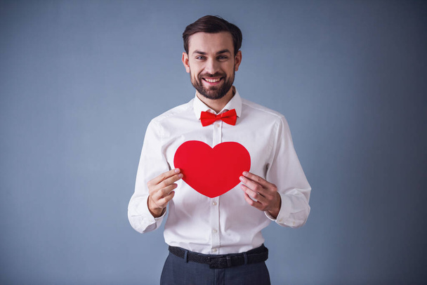 Handsome elegant guy in classic shirt and red bow tie is holding a paper heart, looking at camera and smiling, on gray background - Photo, Image