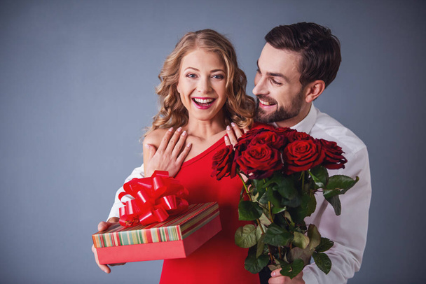 Handsome elegant guy in classic shirt with red bow tie is giving roses and gift box to his beautiful girlfriend and smiling - Photo, Image