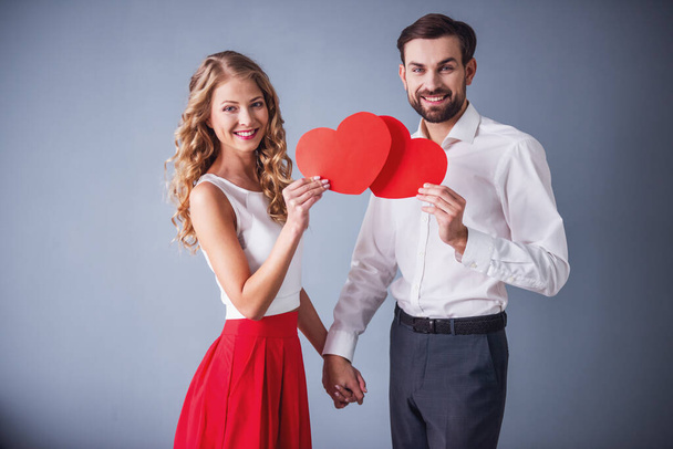 Beautiful elegant couple is holding paper hearts, looking at camera and smiling, on gray background - Photo, Image