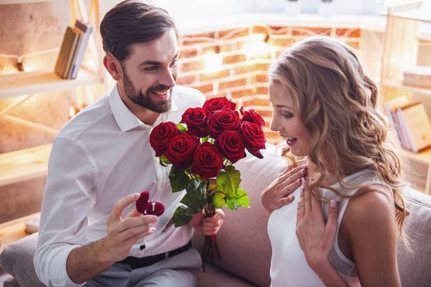 Bonito cara elegante está propondo a sua bela namorada, dando-lhe rosas e sorrindo enquanto eles têm um encontro romântico em casa
 - Foto, Imagem