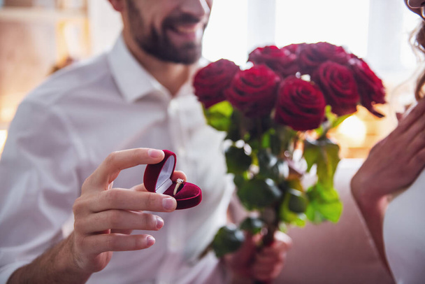 Όμορφος άντρας κομψό προτείνει την όμορφη κοπέλα, δίνοντας της τριαντάφυλλα και χαμογελώντας ενώ έχοντας ένα ρομαντικό ημερομηνία στο σπίτι - Φωτογραφία, εικόνα