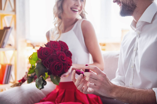 schöner eleganter Kerl macht seiner schönen Freundin einen Heiratsantrag, schenkt ihr Rosen und lächelt, während sie ein romantisches Date zu Hause haben - Foto, Bild