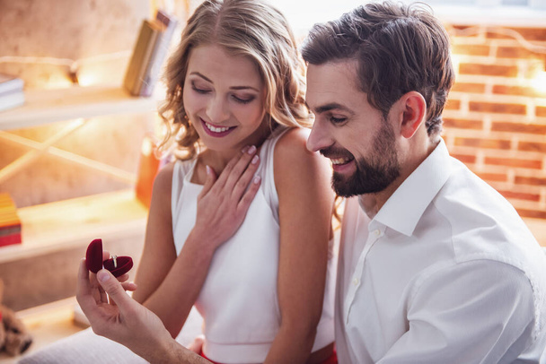 Guapo elegante chico está proponiendo a su hermosa novia y sonriendo mientras tienen una cita romántica en casa
 - Foto, Imagen